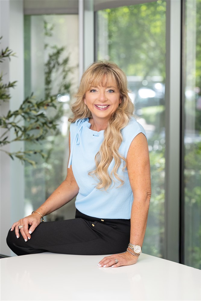 Stacy Harris seated on a table in a bright office wearing a light blue top.